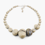 close up of focal hollow blown beads on a flameworked glass necklace. sandstone coloured beads with offset purple-green bead, striped with ivory and blue detail, hand knotted on grey silk cord. shown on white background