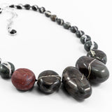 close up of a flameworked glass necklace. hand knotted on black silk cord and showing the fine silver detail on the blown dark charcoal beads and the copper detail on the single etched red offset bead. shown on white background