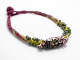 front angle detail of flameworked glass lilac flower headpins strung on a hand knotted and felted necklace in purple with green felt details. on white background.